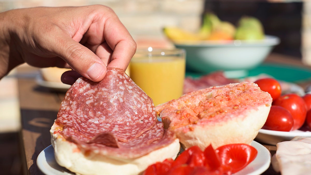 Man's hand seen putting salami on sandwich. Tomatoes, fruit and juice seen in background.
