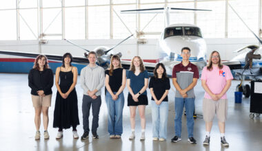 A group of eight people stand together inside a hangar with a somewhat shiny floor. In the background is a small white plane with a blue stripe, and large windows behind that.