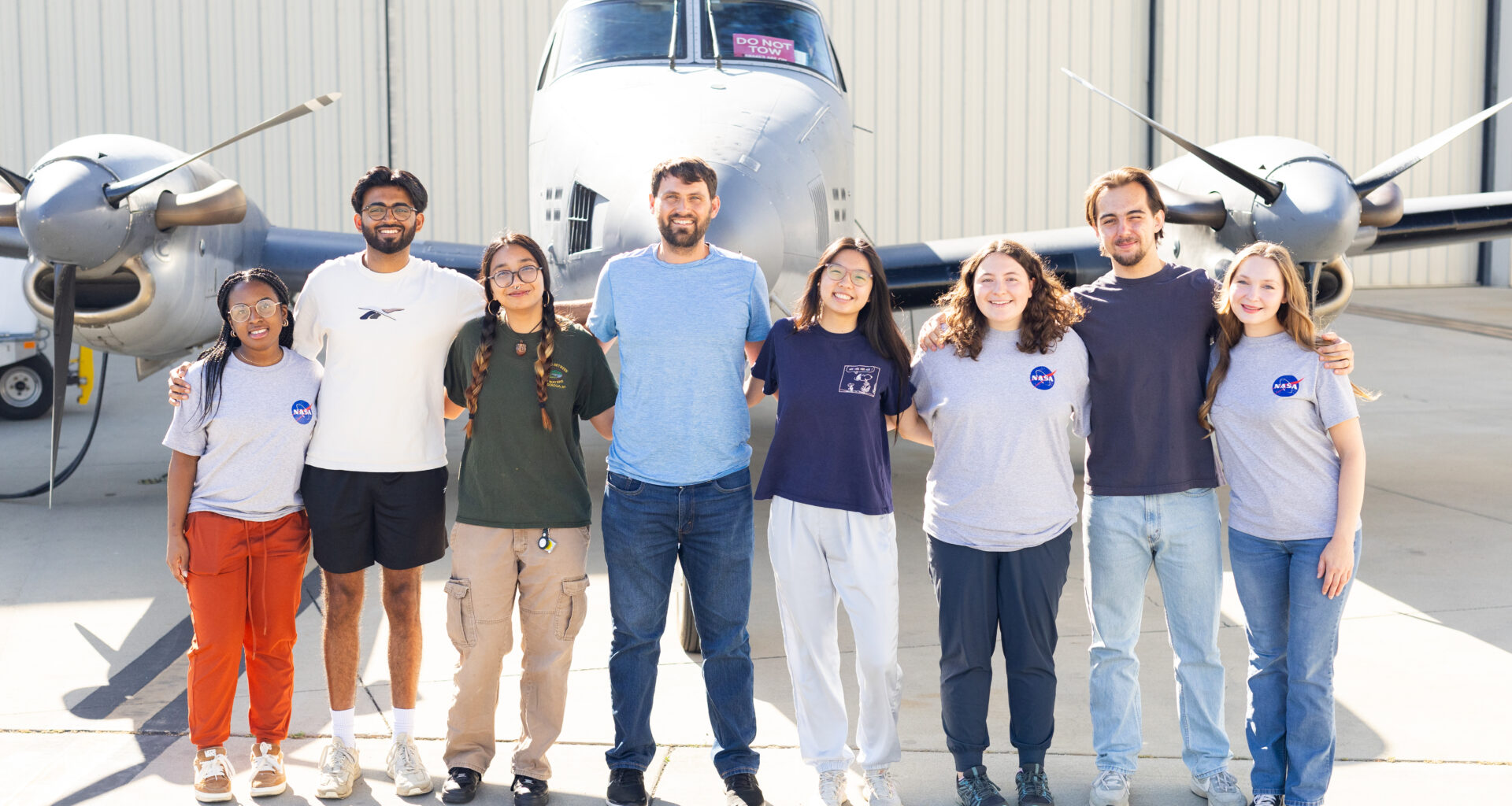 A group of eight people stand on tan tarmac in front of a small gray plane.