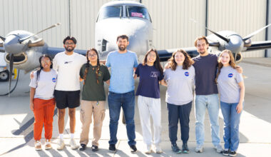A group of eight people stand on tan tarmac in front of a small gray plane.
