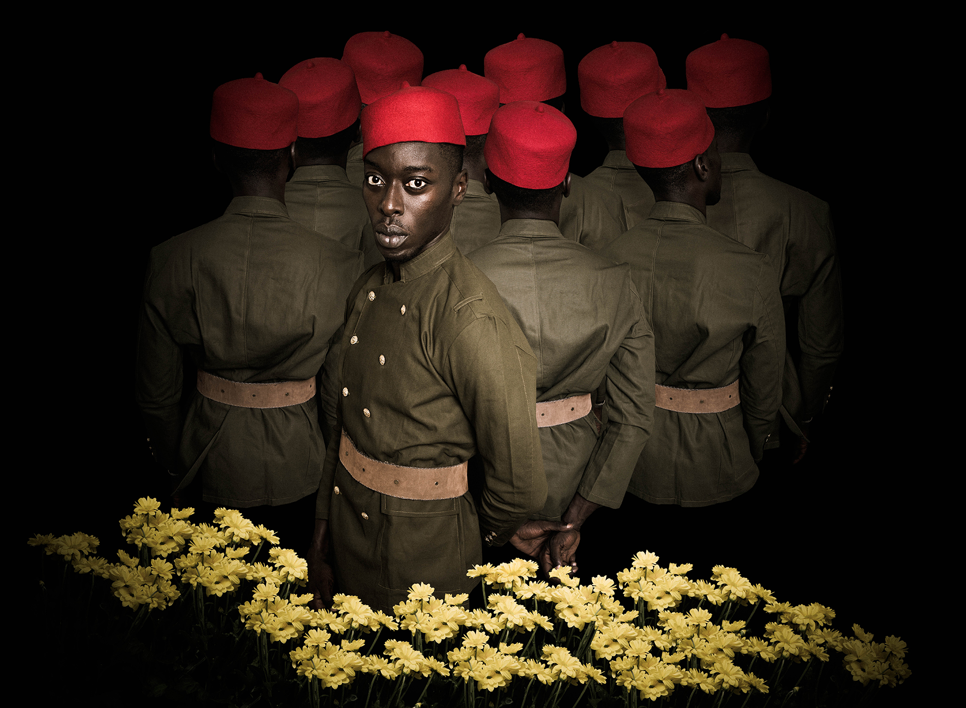 A group of soldiers wearing red caps and green uniforms stands in a line, with yellow flowers blooming in the foreground against a dark background