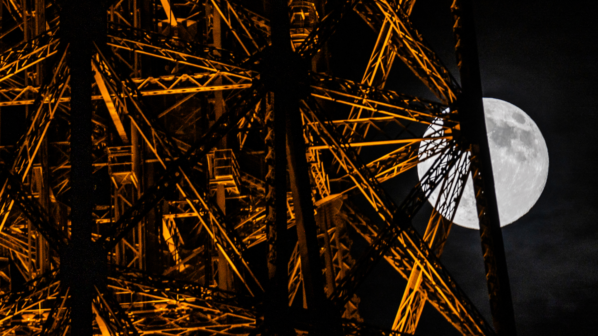 A section of the Eiffel Tower is shown, with the disk of a fully-lit moon emerging from behind its steel girders in a dark sky to the right of the image.