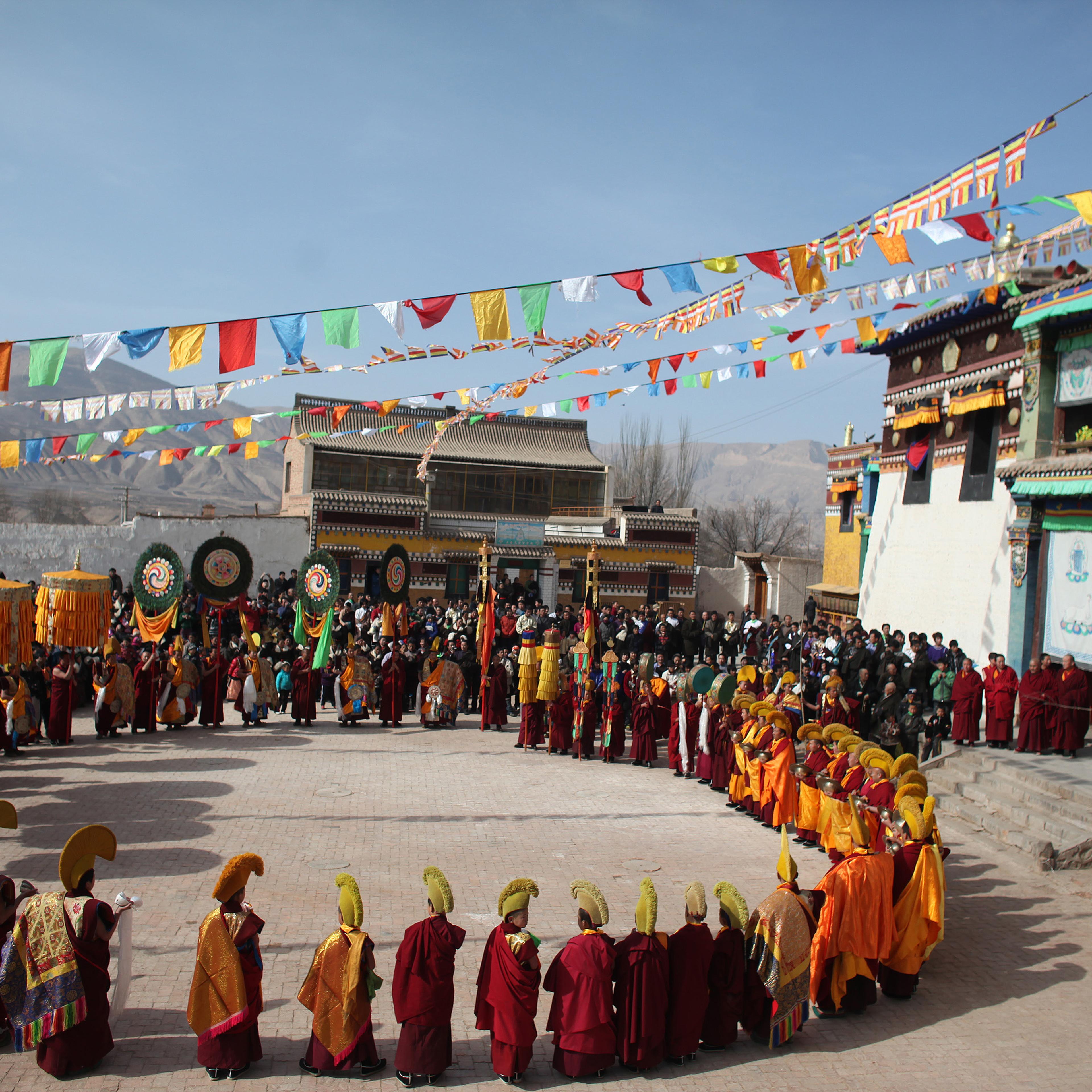 Monks in colourful robes and hats, forming a circle during an outdoor festival with flags and onlookers in Tibet.