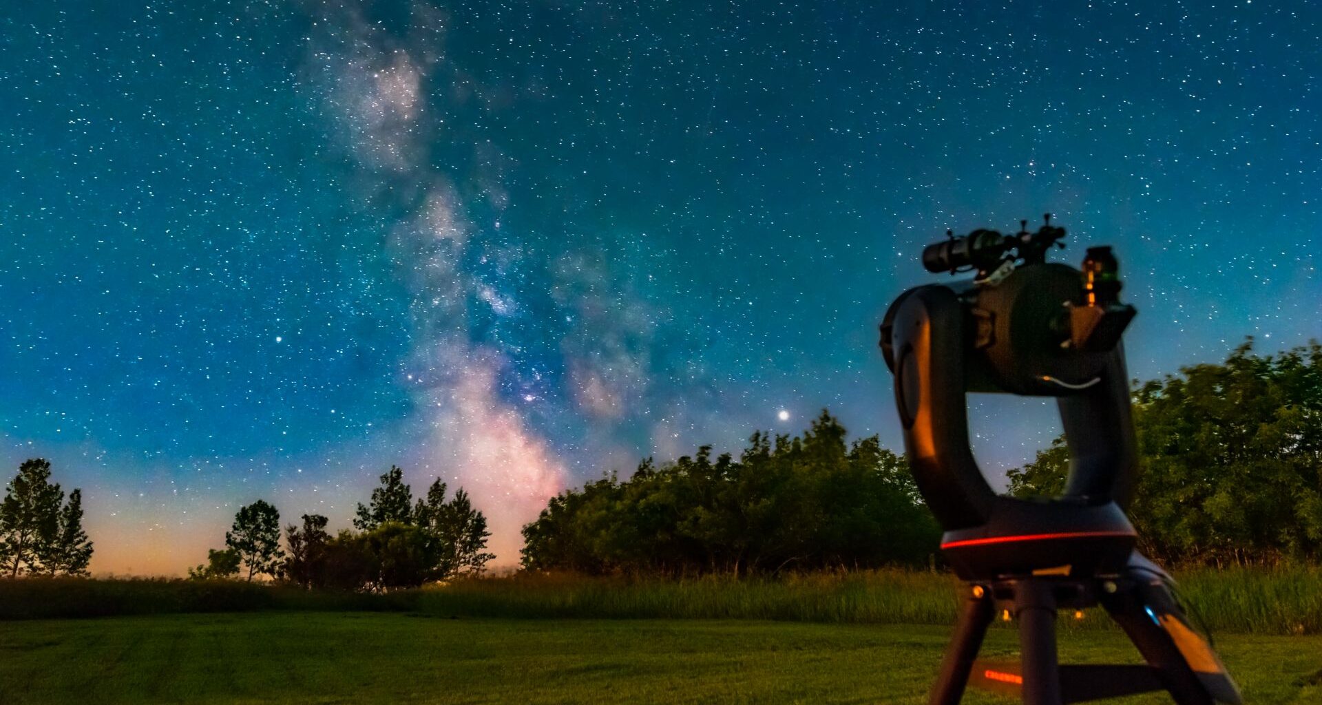 A telescope on a tripod sits in a grassy field looking at the pink glow of the Milky Way galaxy past some trees with a light blue night sky behind