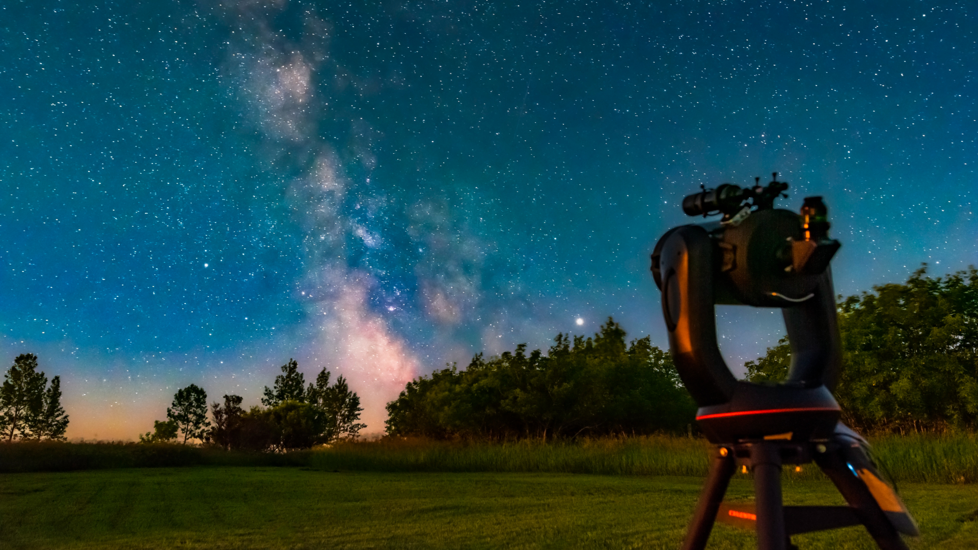 A telescope is pictured to the right, looking out over a rural field as the Milky Way shines overhead in the night sky.
