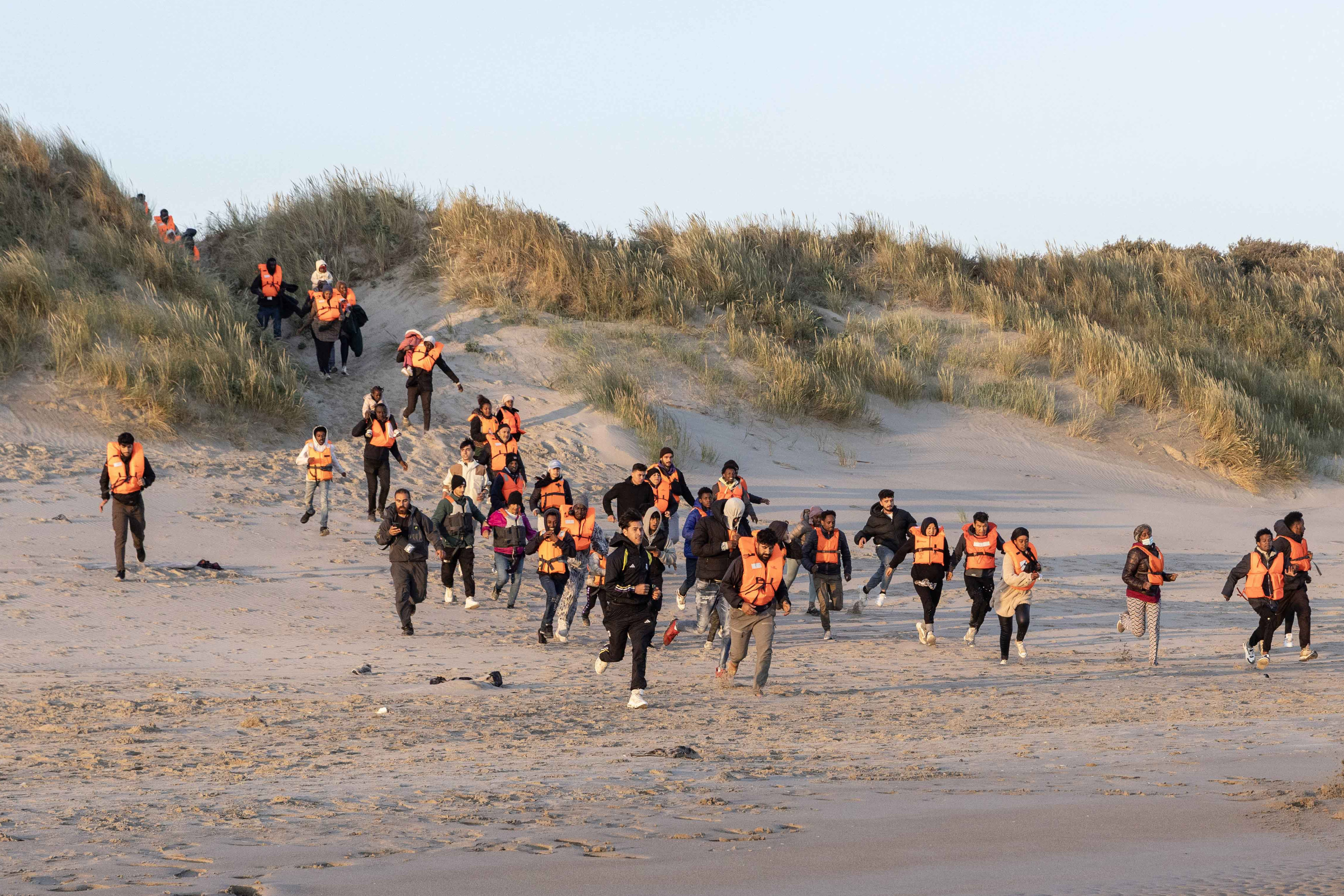 Migrants wearing life vests run from sand dunes towards a beach.