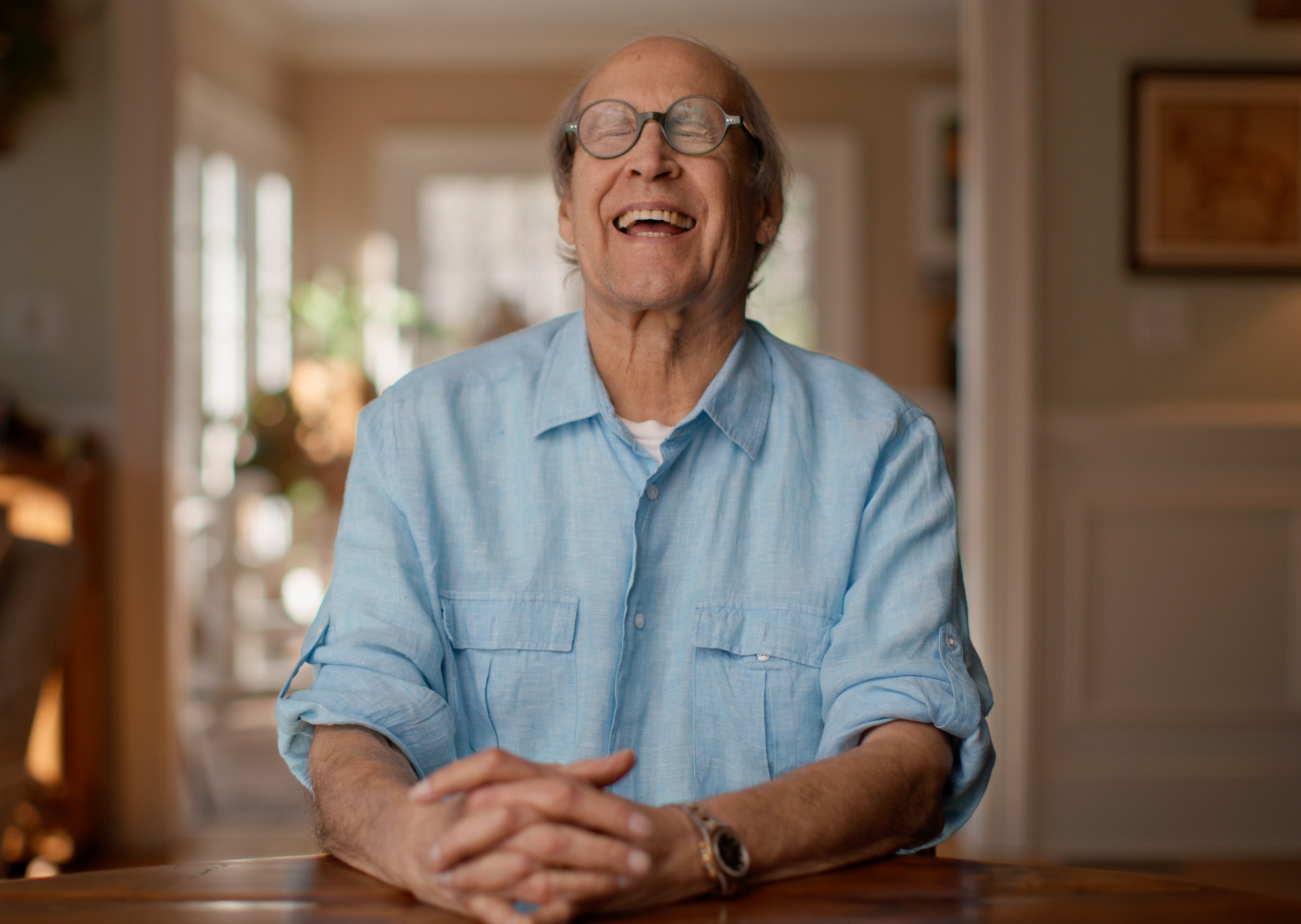 Chevy Chase in a light blue shirt and round glasses, laughing with his hands clasped on a table.