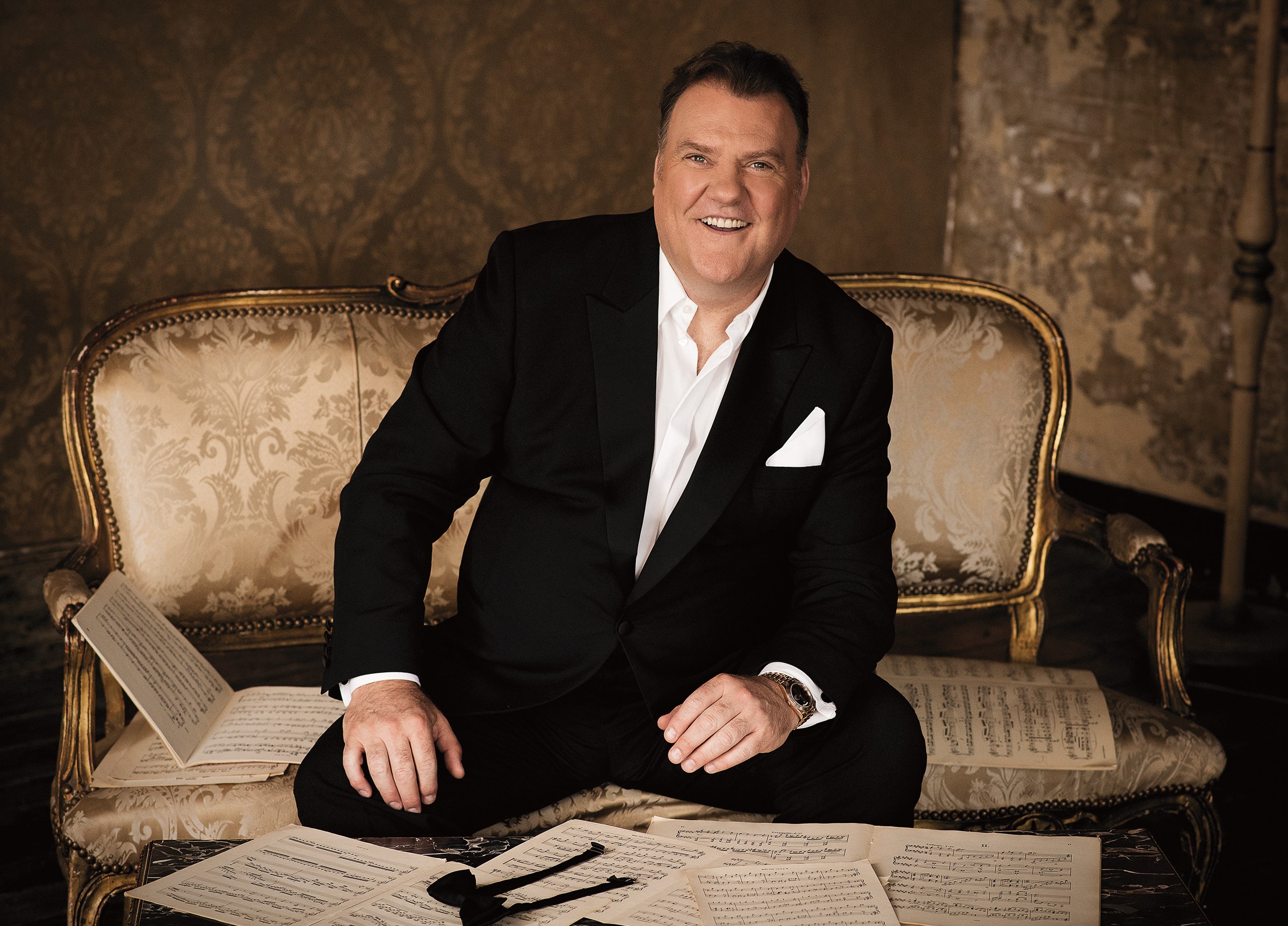 Portrait of Bryn Terfel smiling while sitting on a patterned sofa with sheet music.