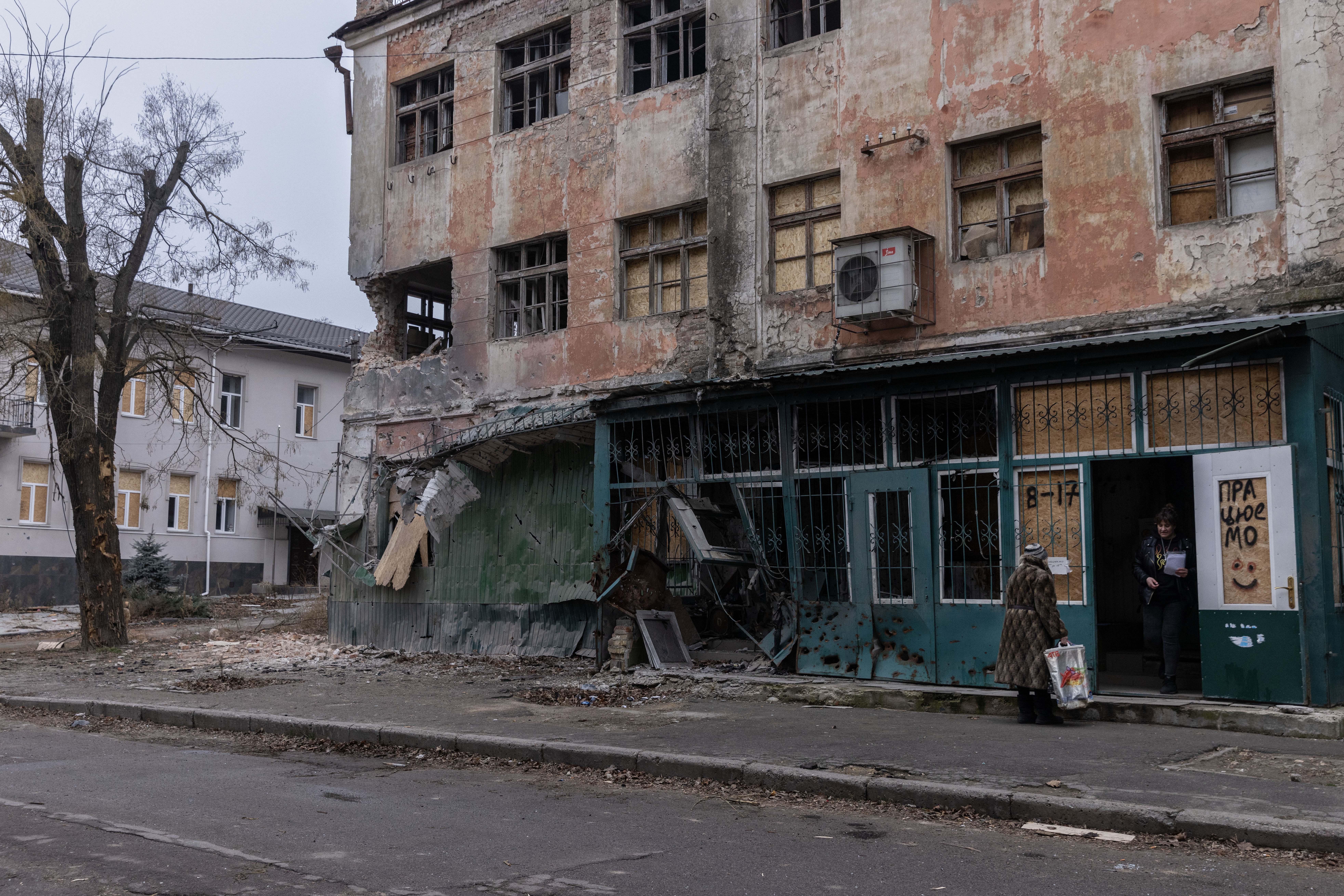 A war-damaged building with peeling paint and boarded-up windows, with a woman standing in the doorway of an open shop.