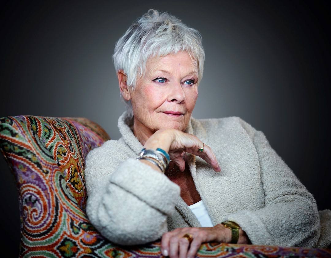 Dame Judi Dench seated in a colorful patterned armchair.