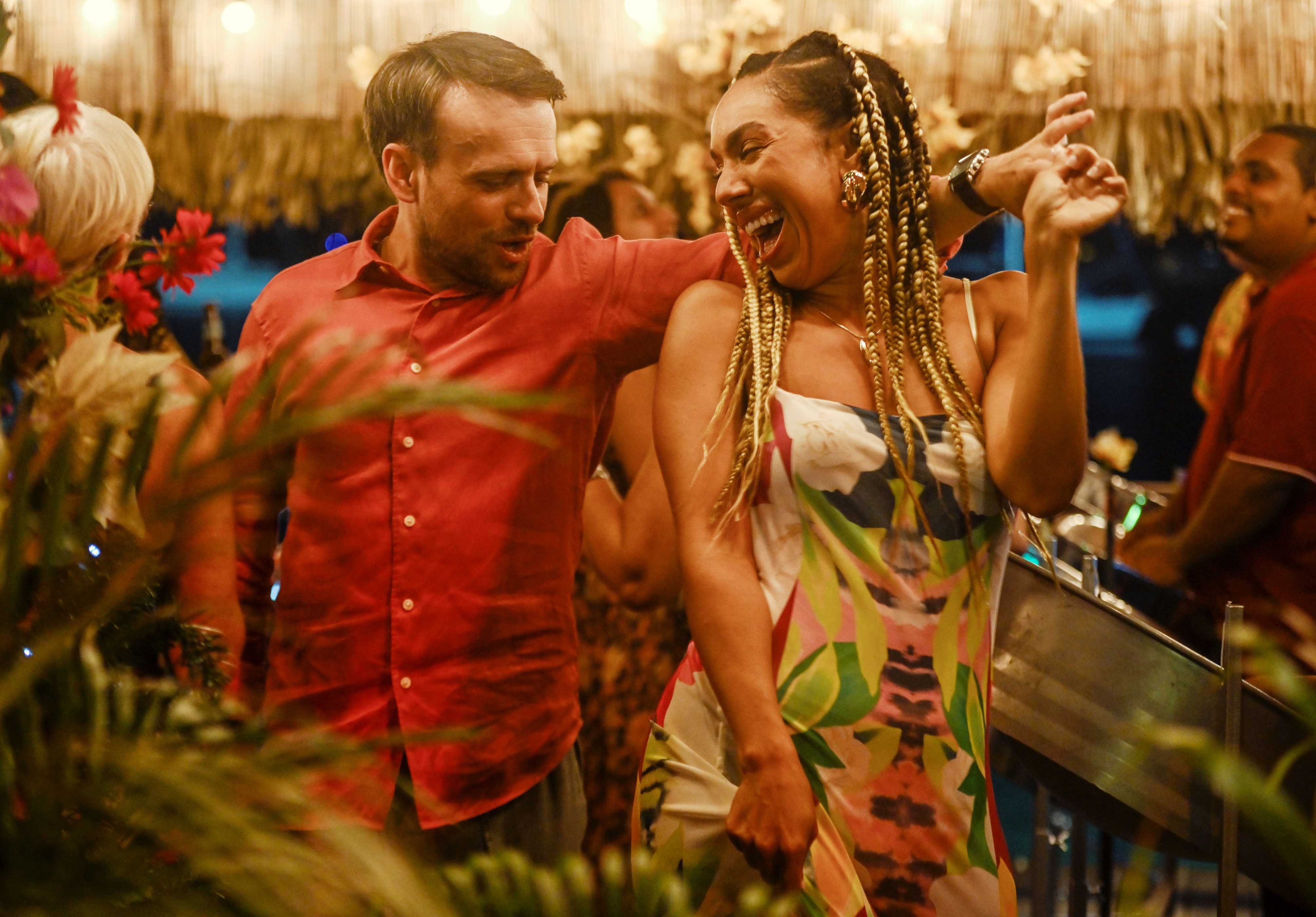 A man in a red shirt with his arm around a laughing woman in a patterned dress and braided hair.