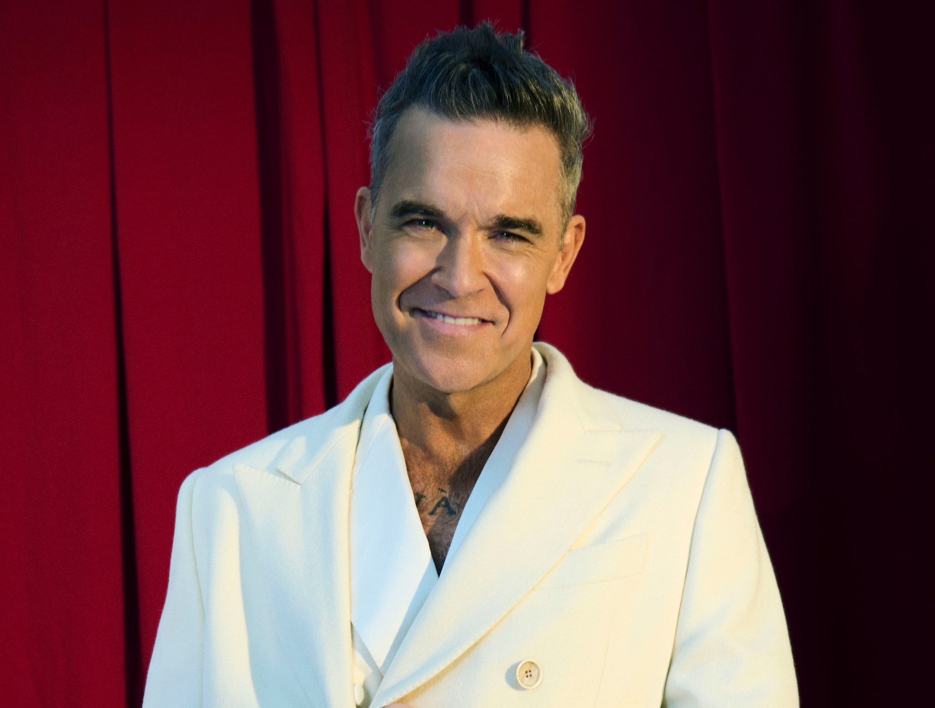Robbie Williams in a white suit, standing in front of a red curtain.