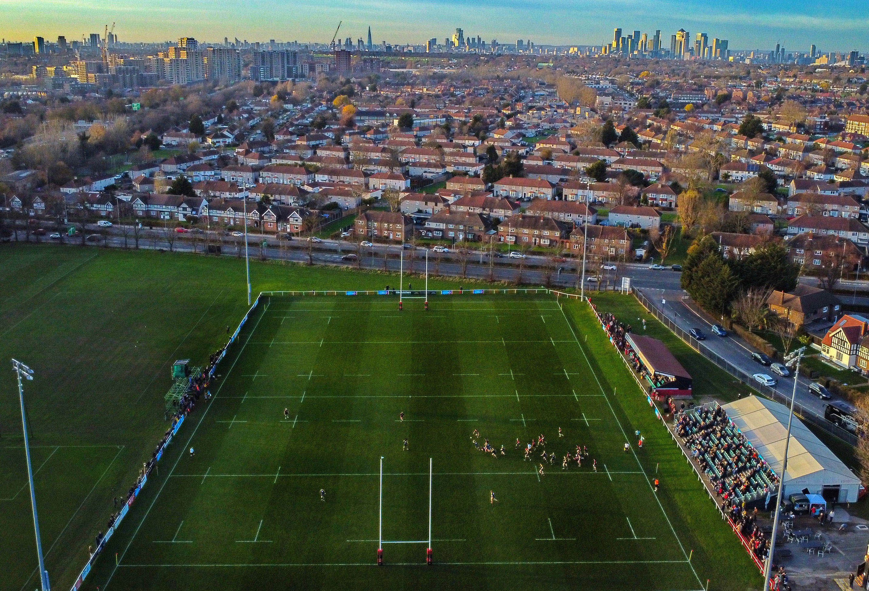Aerial view of a rugby game at Blackheath with players and spectators on the field and buildings and cityscape in the background.