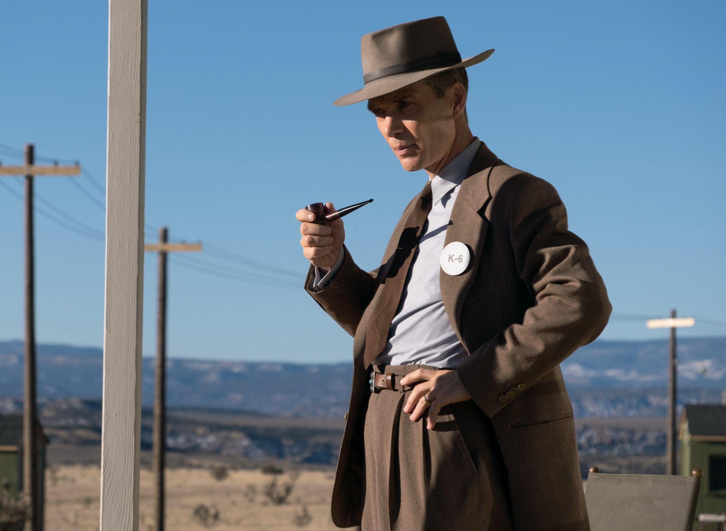 Cillian Murphy as Oppenheimer, wearing a hat, brown suit, and an identification badge, holding a pipe, with a desert landscape in the background.