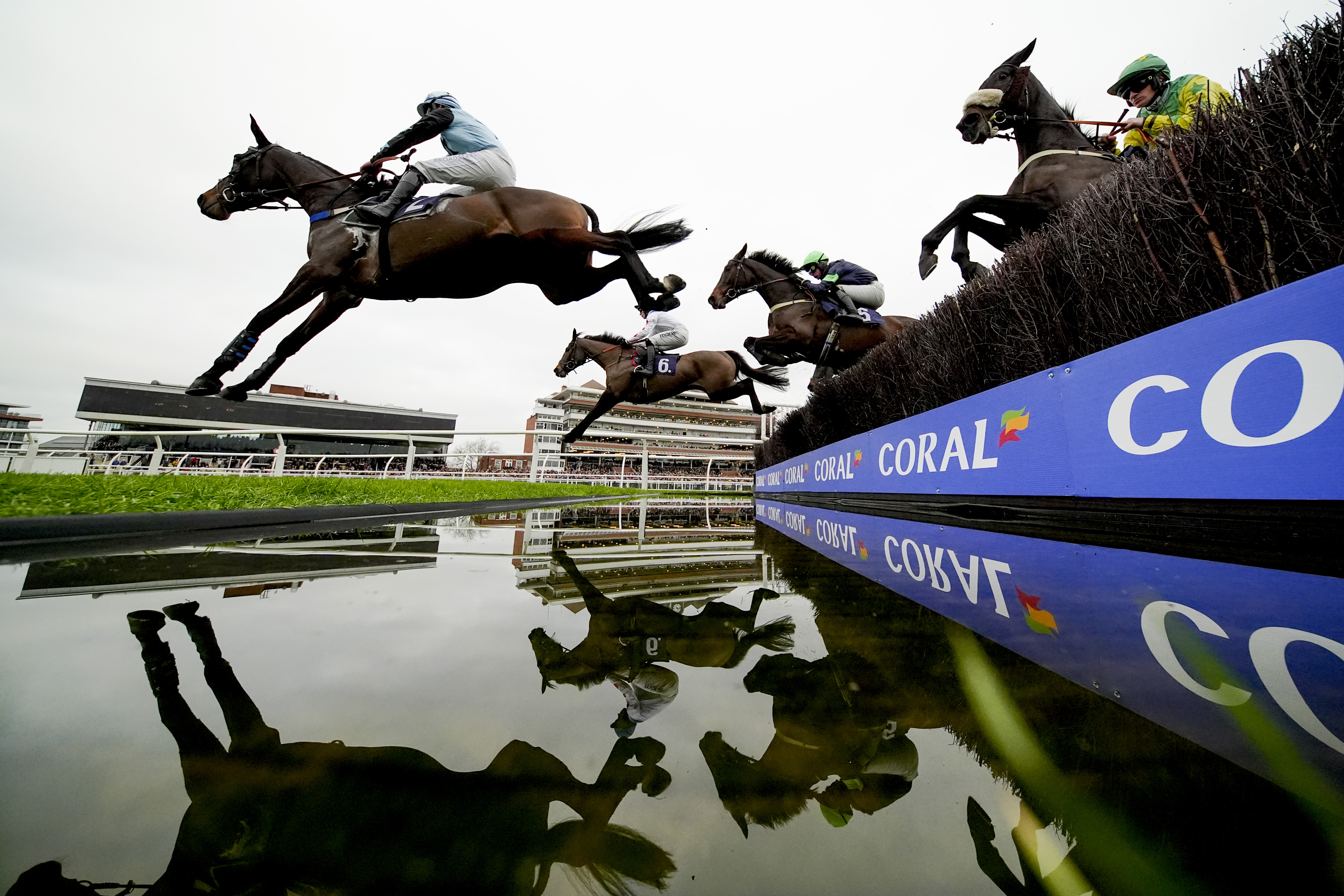 Horses and jockeys jumping over a water obstacle at Newbury Racecourse.