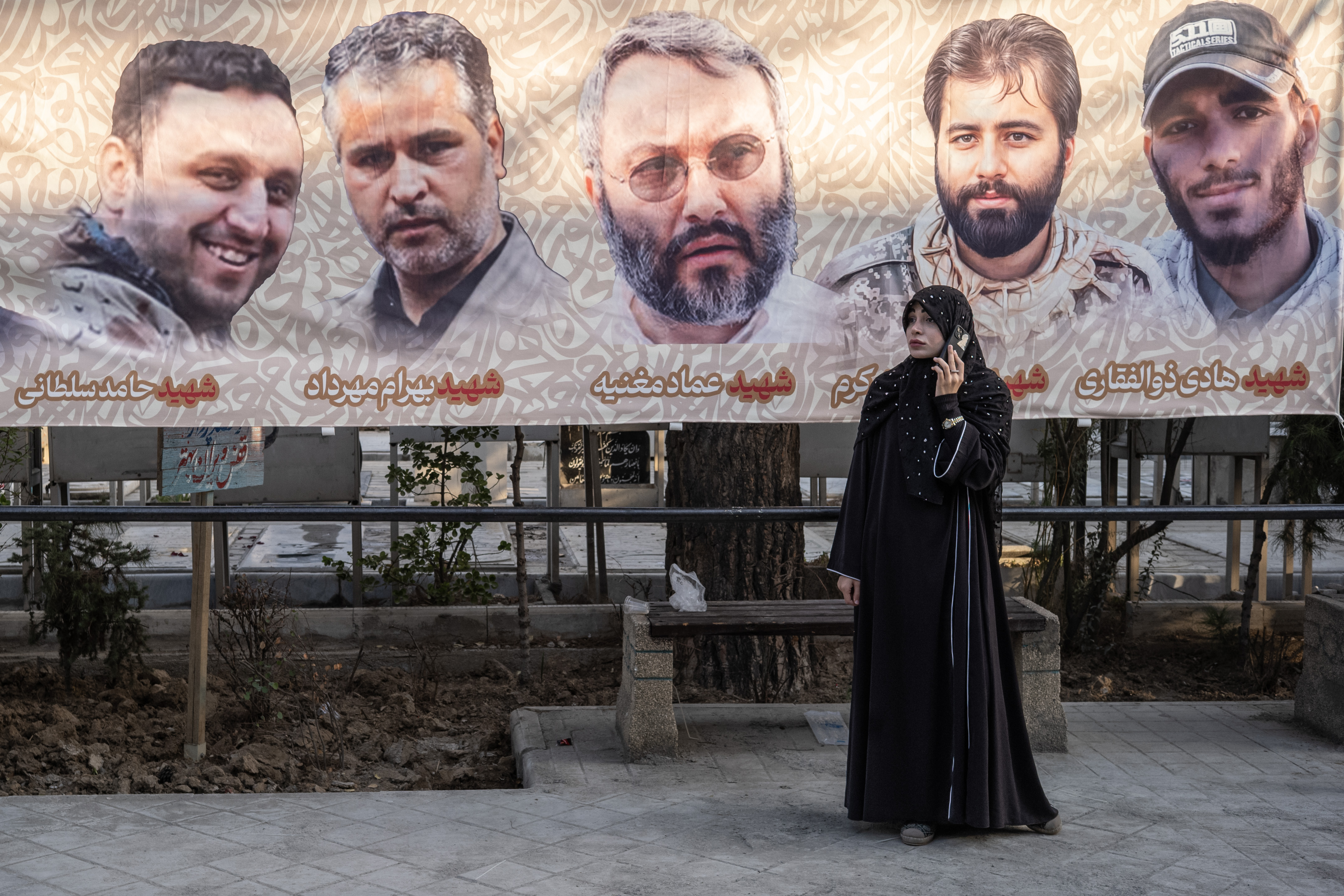 Woman on cell phone standing in front of a banner of five men at the Martyr's section of Behesht-e Zahra cemetery.