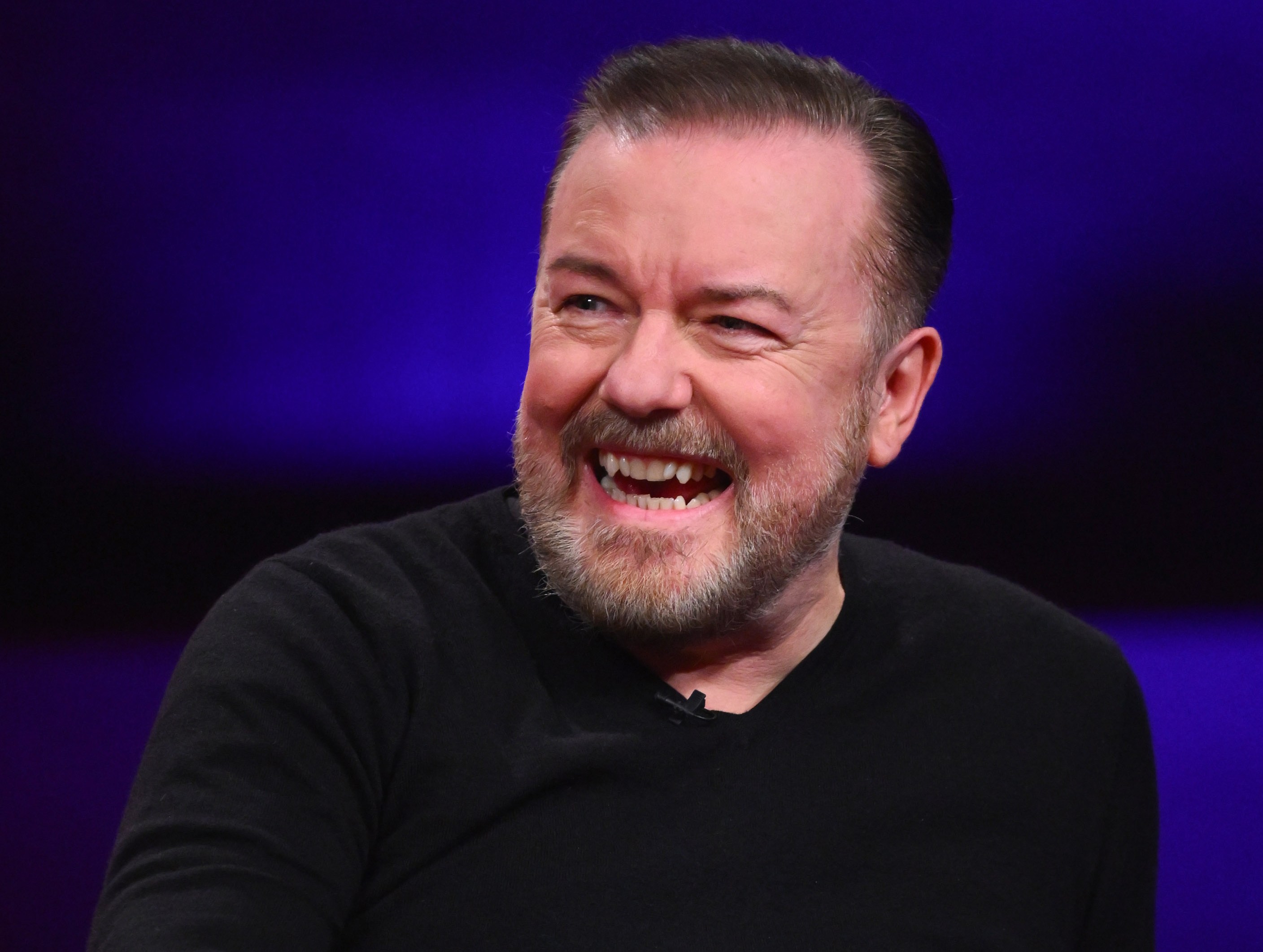 Ricky Gervais laughing on a red couch.