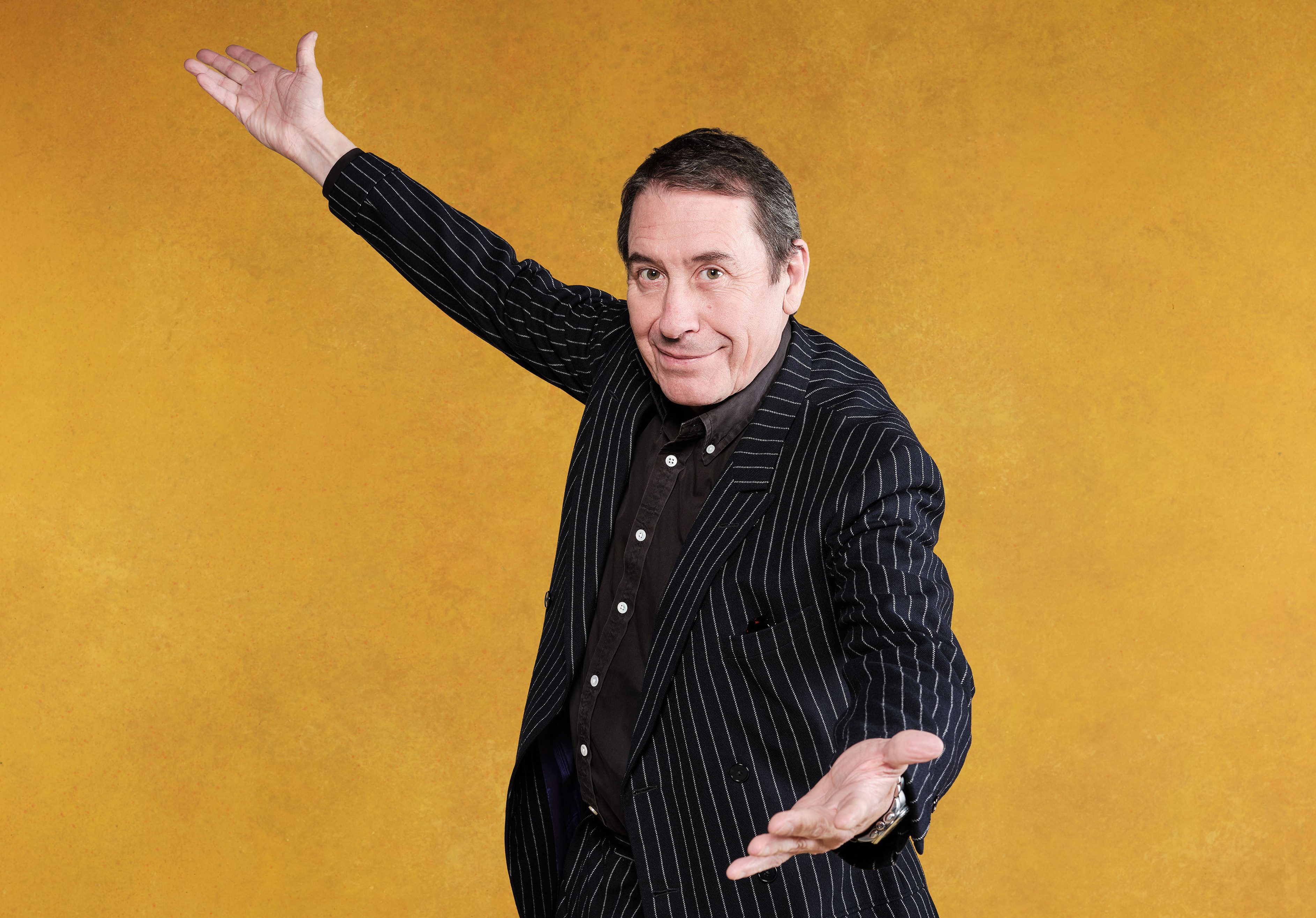 Jools Holland in a black pinstripe suit and black shirt, with his arms outstretched as if presenting.