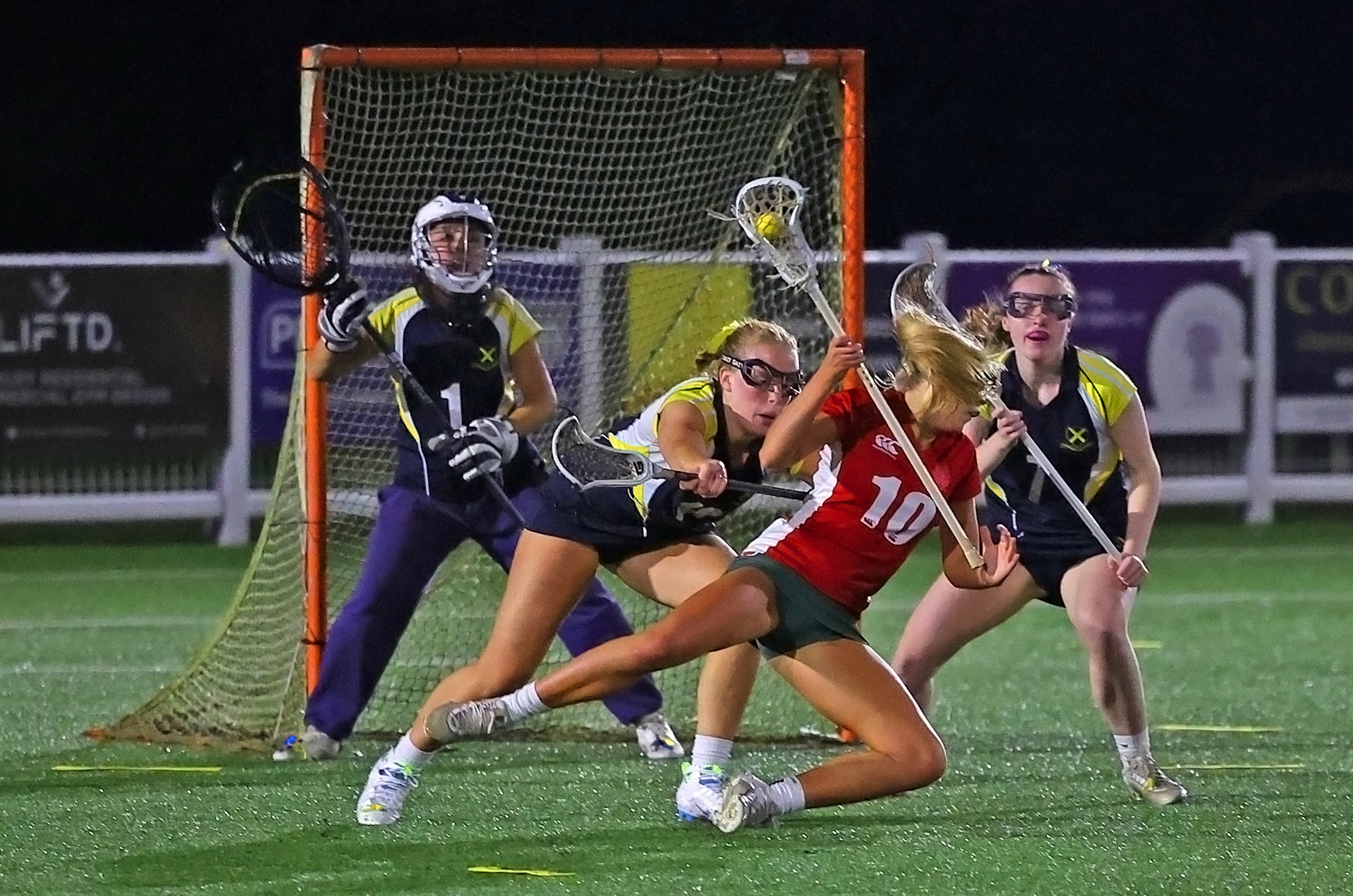 Four female lacrosse players on an artificial turf field at night, with one player in red attempting a shot while two defenders in black and yellow and a goalie in purple try to block her.