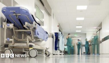 A generic image of a hospital corridor, with a stretcher on the left, and staff wearing green surgical uniforms, with blurred faces, in the distance.