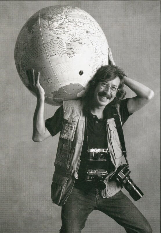 A smiling man with glasses holds a large globe on his shoulder. He wears a photography vest with a "PRESS" patch and has two cameras hanging around his neck. The background is plain and light-colored.
