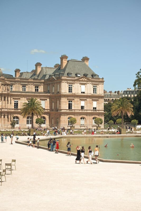 People walk and gather around a large pond with small sailboats in front of the Luxembourg Palace, a historic building with classic French architecture, surrounded by palm trees and greenery on a sunny day.