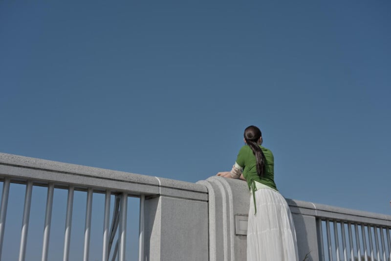A woman with long dark hair, wearing a green top and long white skirt, leans on a concrete bridge railing, looking up at the clear blue sky.