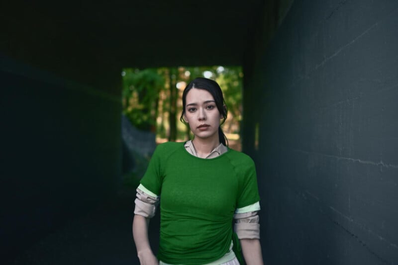A young woman with long dark hair, wearing a green shirt over a light long-sleeve, stands in a dimly lit tunnel with sunlight and green trees visible in the background. She looks directly at the camera with a neutral expression.