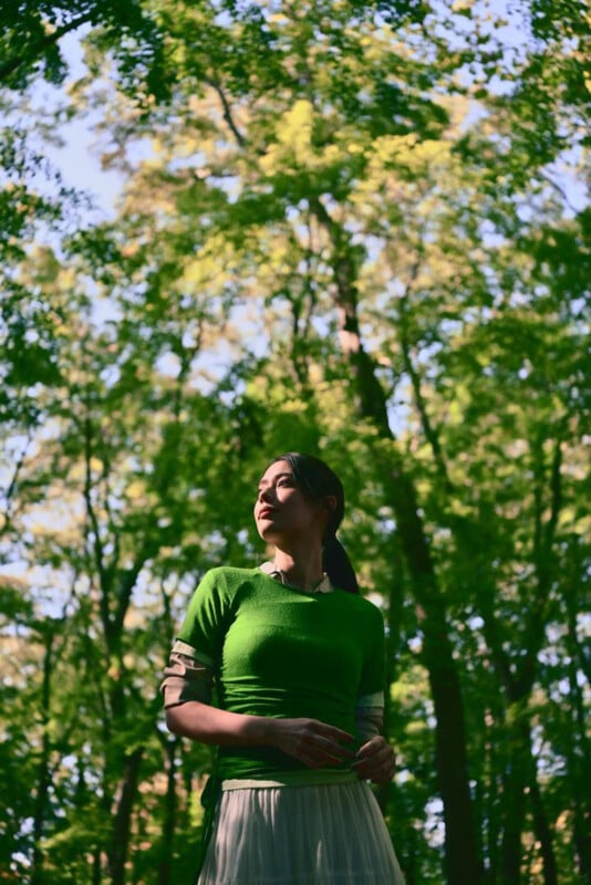 A woman in a green top and white skirt stands in a sunlit forest, looking to the side, with tall trees and green foliage surrounding her. Dappled sunlight creates patterns on her and the forest floor.