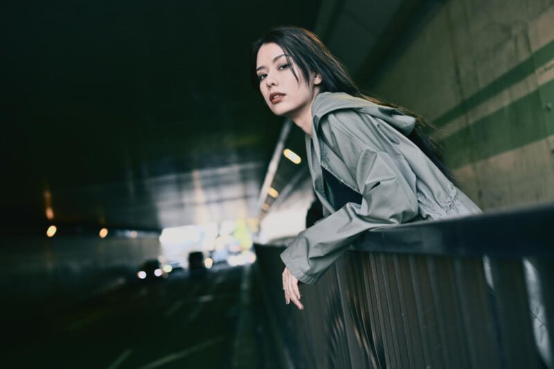 A young woman in a gray-green jacket leans on a railing in a dimly lit tunnel, looking at the camera with a serious expression. Blurred cars and lights are visible in the background.