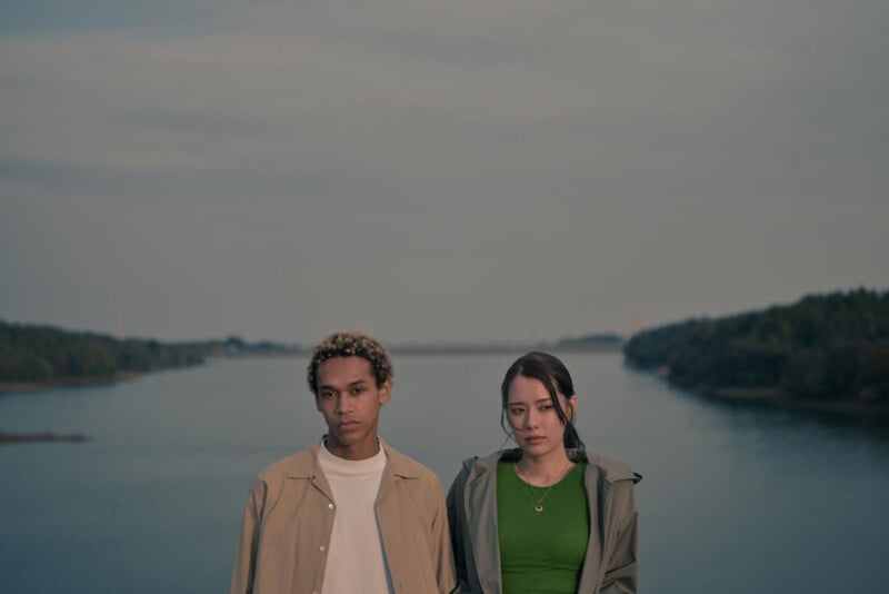 Two people stand side by side in front of a calm river, with trees lining both sides under a cloudy sky. They both have neutral expressions and wear earth-toned clothing.