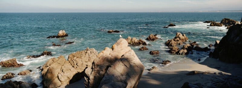 Rocky coastline with large boulders and waves crashing against them, bordered by clear blue ocean water under a bright sky, with sandy beach areas visible between the rocks.