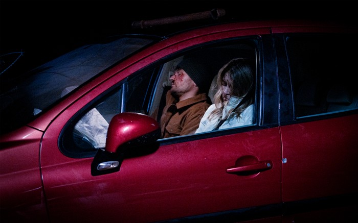 Charity and Mack, covered in blood, lie unconscious in a red car in a scene from Corriedale