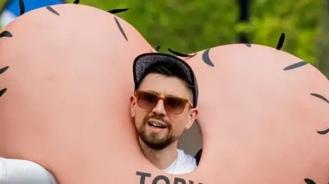 Robin Cancer Trust Toby Freeman during a run. He is wearing sunglasses and a cap and is wearing an outfit designed to look like a pair of large testicles.