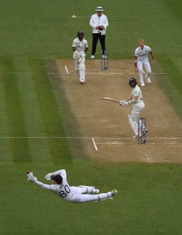 A cricket wicketkeeper dives to his left to attempt a catch, while the batsman stands at the crease and the bowler and umpire look on during a match.