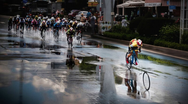 A group of cyclists races on a wet road, with one cyclist ahead who has lost control as their front wheel detaches, while the other competitors follow behind, splashing through water.