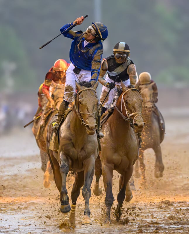 Jockeys riding muddy horses race on a wet, dirt track. The leading jockey in blue raises a whip while another closely follows, with mud flying from the horses’ hooves. Other racers trail behind in the background.