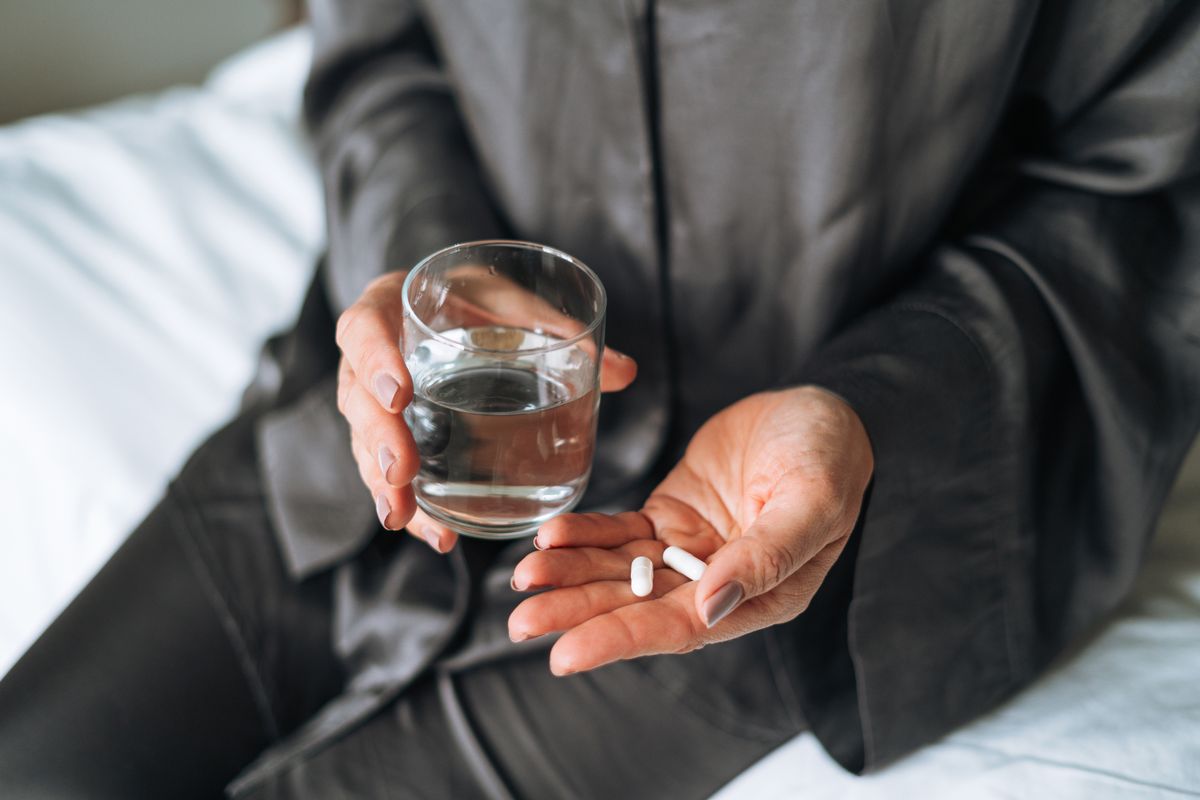Woman in pajamas drinking water with pills sitting on her bed at home