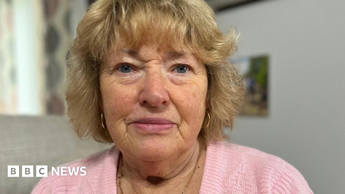 Joyce Gifford with blue eyes and golden-coloured, short, wavy hair with a fringe. She is wearing a baby pink wool jumper with a thin, gold necklace and thin, gold, hoop earrings.