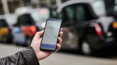 Getty Images A hand holding a mobile phone which is on a map app. The arm shows the person is wearing a black leather jacket and behind their phone is a line of black taxis.