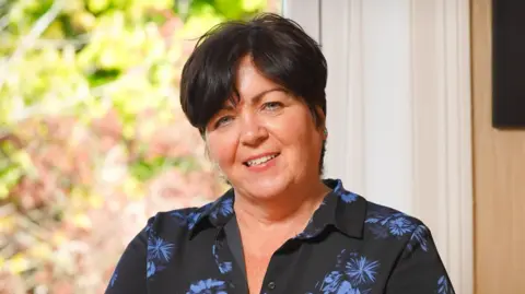 Royal College of Nursing A professional headshot of Rita Devlin. She has short, dark hair and is smiling at the camera. She is wearing a dark shirt with blue flower patterns. She is sitting in front of the window.