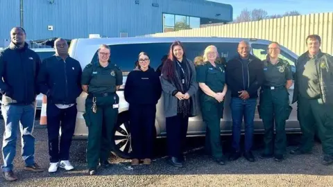 East of England Ambulance Service A group of nine staff, including paramedics stood together outside a large silver car with darkened windows.