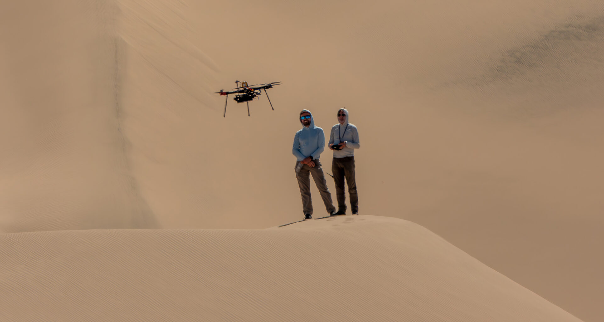 Two people stand side by side on a sandy hill, or dune. The person on the left is standing in a blue top, while the person on the right in a gray top is holding a controller. Above and to the left of their heads is a rotorcraft flying above the dune. The background of this image is more sandy dunes.