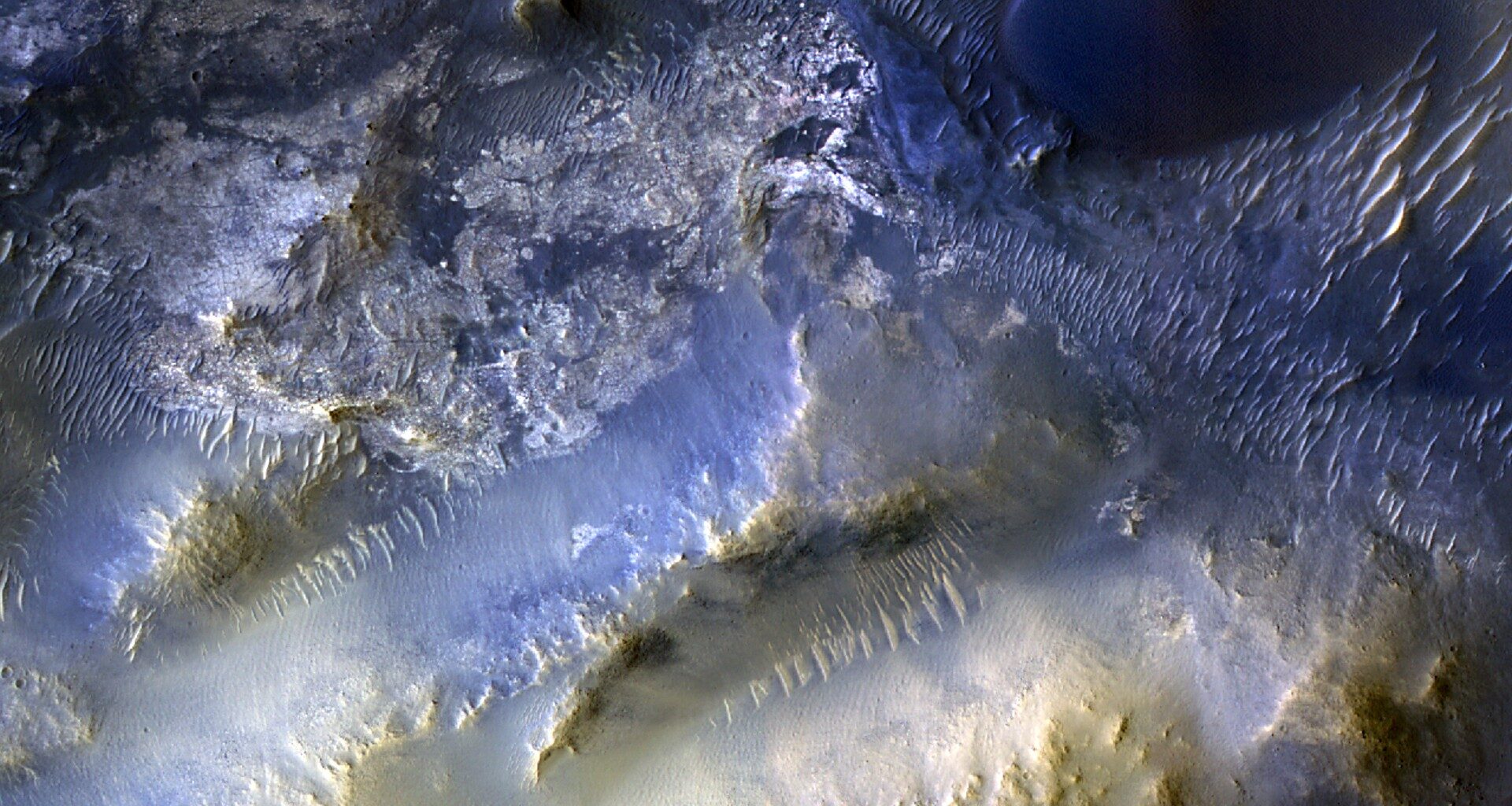 An overhead color view of the Martian surface shows rough and varied terrain in shades ranging from golden tan to electric blue. At upper left, the ground appears more flat and cratered than other areas of the image, and is colored dark grayish-blue with highlights of silver and tan. The upper right corner of the image looks like wavy sand dunes, in shades of dark blue with bright silvery highlights, except for one apparent ridgeline that stands out as an elongated S-shape in electric blue. The bottom half of the image shows more mountainous terrain that gets lighter and more gold-colored nearer the bottom of the frame. A pair of smooth valleys run diagonally between the peaks, from around the center of the image toward the bottom-left corner; the upper one is a shade of silvery blue and the bottom one is a grayish-gold, and both have ridges lining their upper walls, looking like lines of sharp teeth biting into the valleys.