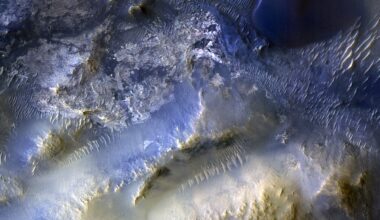 An overhead color view of the Martian surface shows rough and varied terrain in shades ranging from golden tan to electric blue. At upper left, the ground appears more flat and cratered than other areas of the image, and is colored dark grayish-blue with highlights of silver and tan. The upper right corner of the image looks like wavy sand dunes, in shades of dark blue with bright silvery highlights, except for one apparent ridgeline that stands out as an elongated S-shape in electric blue. The bottom half of the image shows more mountainous terrain that gets lighter and more gold-colored nearer the bottom of the frame. A pair of smooth valleys run diagonally between the peaks, from around the center of the image toward the bottom-left corner; the upper one is a shade of silvery blue and the bottom one is a grayish-gold, and both have ridges lining their upper walls, looking like lines of sharp teeth biting into the valleys.