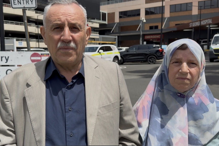 A grey haired man and a woman wearing a hijab stand outside a hospital where their son is recovering.