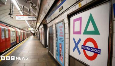 Taken on the Tube platform, a red and white train is seen moving away from the camera - with an empty platform. On the right in the foreground is a Tube sign saying Oxford Circus and other adverts are seen going into the distance.