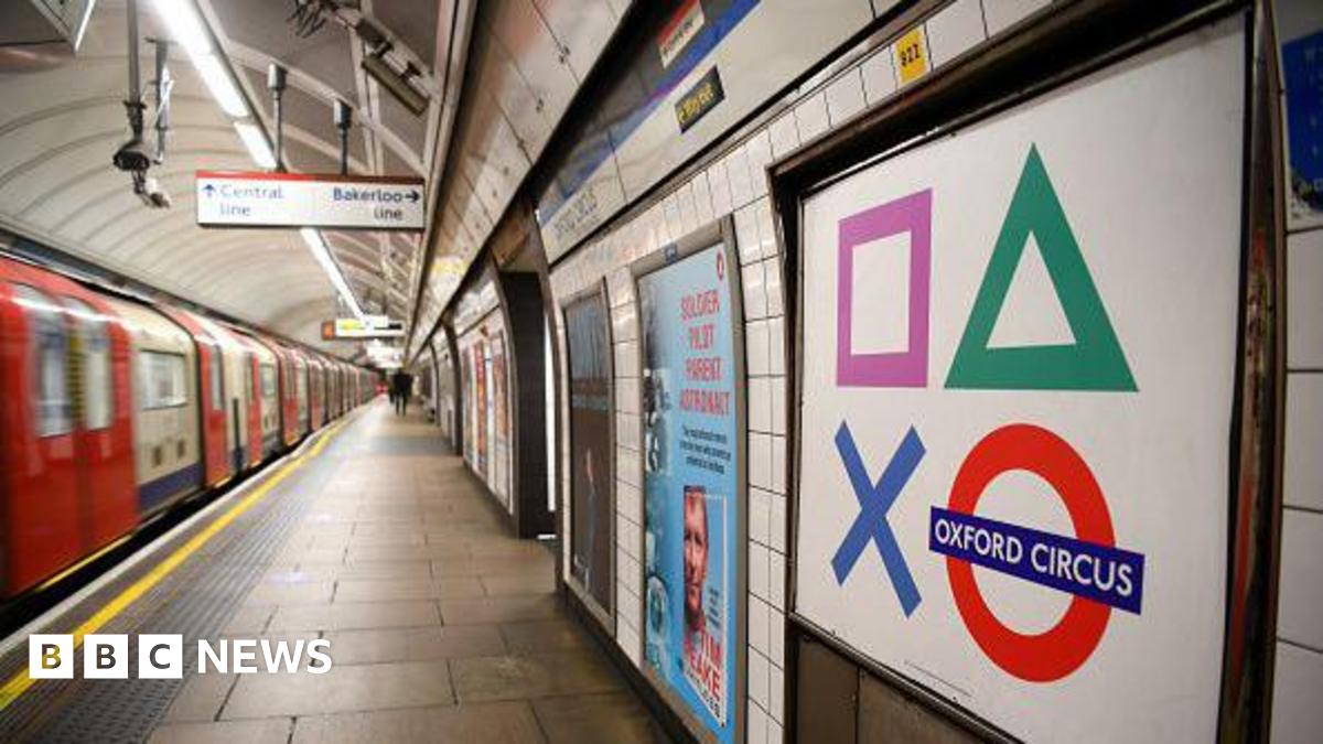 Taken on the Tube platform, a red and white train is seen moving away from the camera - with an empty platform. On the right in the foreground is a Tube sign saying Oxford Circus and other adverts are seen going into the distance.