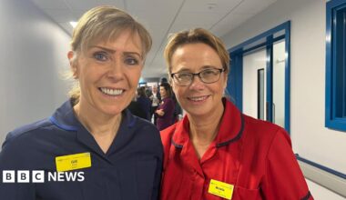 Gill and Michelle stood side by side, smiling and looking into the camera. Gill, on the left, is wearing a dark blue nurse's uniform and a yellow name badge. Michelle is wearing a bright red nurse's uniform and a yellow name badge too. They're stood in a long, white corridor with blue window and door frames.
