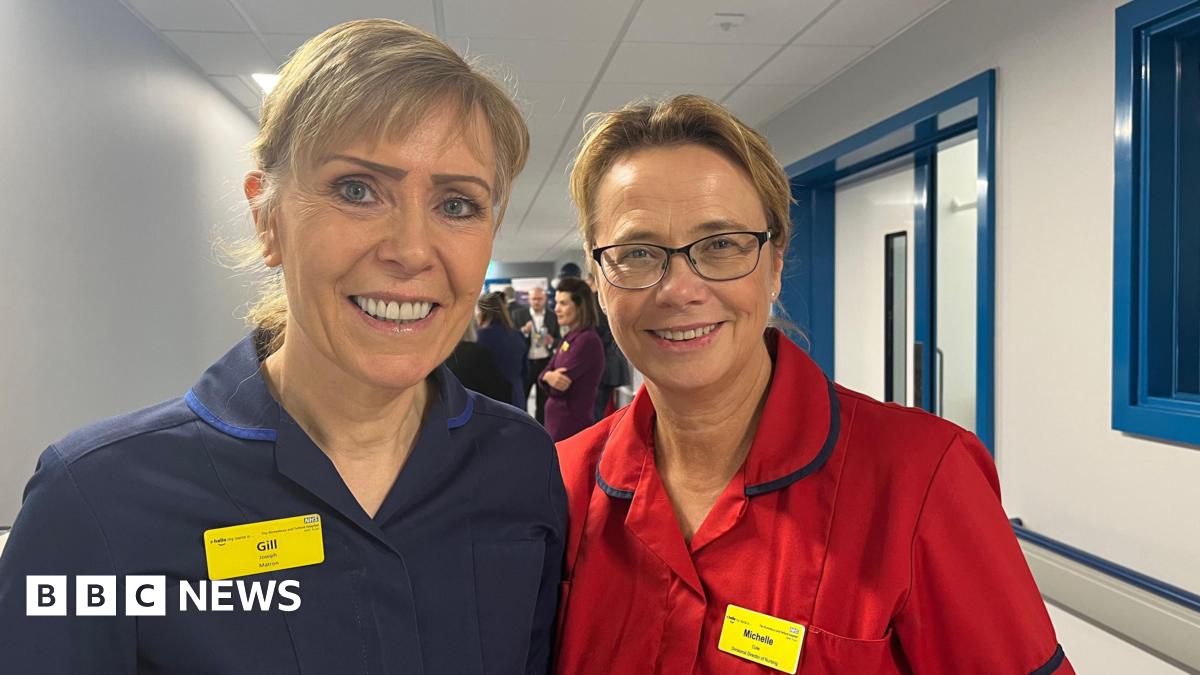 Gill and Michelle stood side by side, smiling and looking into the camera. Gill, on the left, is wearing a dark blue nurse's uniform and a yellow name badge. Michelle is wearing a bright red nurse's uniform and a yellow name badge too. They're stood in a long, white corridor with blue window and door frames.